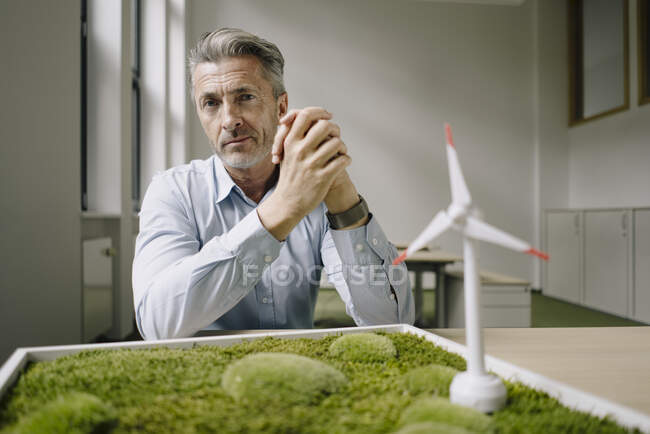 Empresario con las manos apretadas sentado por el juguete de la turbina eólica y el marco de musgo en la mesa en la oficina - foto de stock