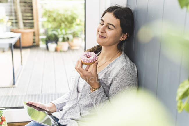 Woman with eyes closed holding digital tablet and doughnut while sitting at home — Stock Photo