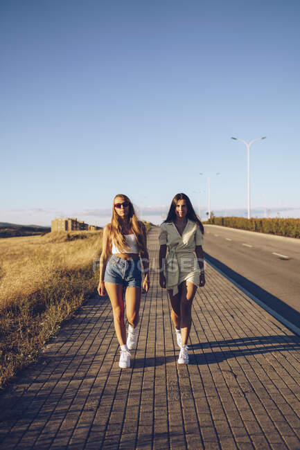 Молоді друзі-жінки, що ходять на тротуарі проти неба в сонячний день — стокове фото