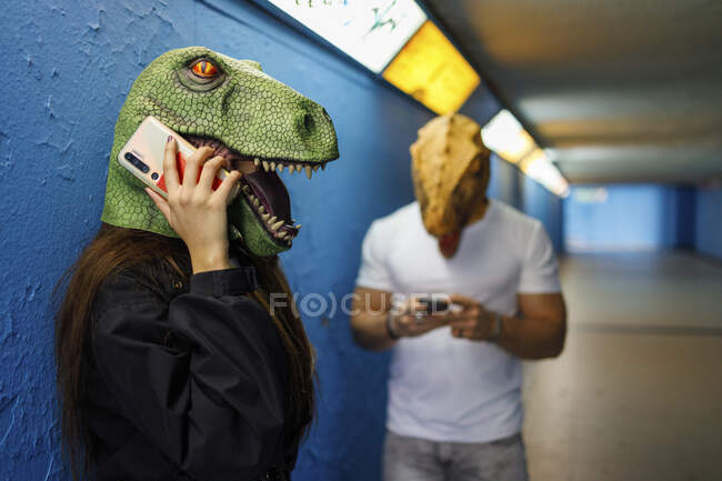 Mujer hablando en el teléfono inteligente por un amigo varón mientras usa máscara de dinosaurio contra la pared azul - foto de stock