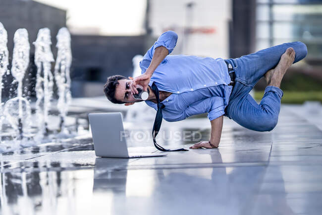 Young businessman with laptop drinking coffee while doing handstand by fountain on footpath — Stock Photo