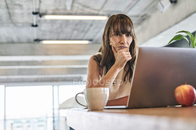 Mujer reflexiva con la mano en la barbilla usando el ordenador portátil mientras está sentado en casa - foto de stock