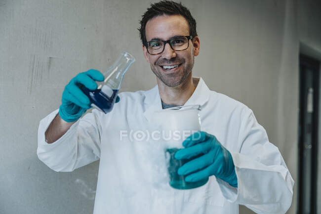 Hombre sonriente sosteniendo un vaso de precipitados de solución líquida mientras está parado en el pasillo de la clínica - foto de stock