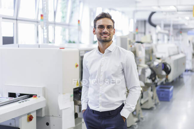 Lächelnder junger Geschäftsmann steht mit Händen in den Taschen vor beleuchteter Fabrik — Stockfoto