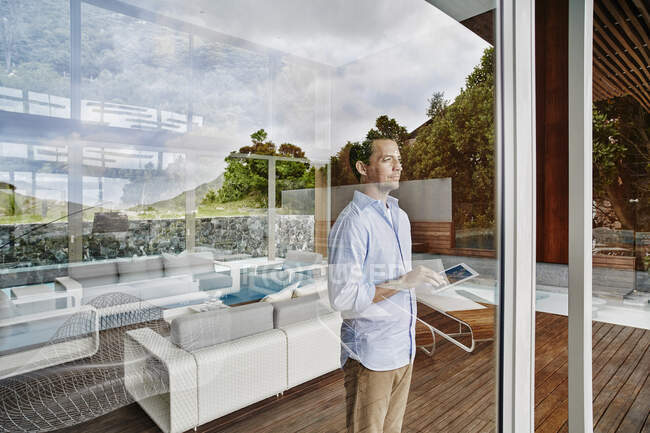 Зрелый человек, использующий цифровой планшет, глядя в окно, стоя дома — стоковое фото