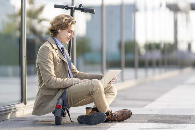 Empresario que usa auriculares en la oreja usando un portátil mientras está sentado en un scooter eléctrico - foto de stock