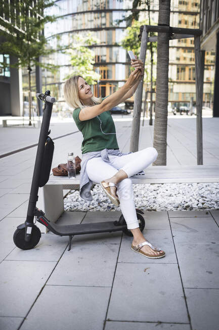 Mitte erwachsene Frau hört Musik und macht Selfie, während sie auf Bank in der Stadt sitzt — Stockfoto