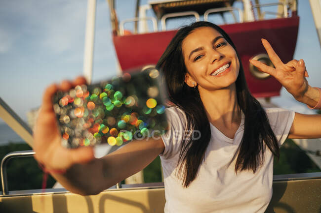 Beautiful young woman taking selfie through smart phone while gesturing peace sign on Ferris wheel — Stock Photo