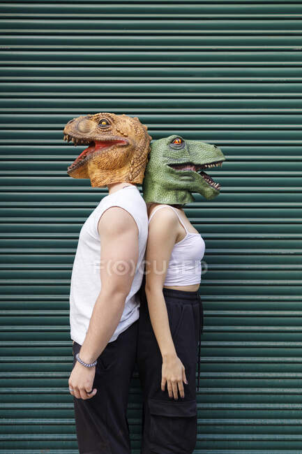 Amigos hombres y mujeres en máscara de dinosaurio de pie contra el obturador verde - foto de stock