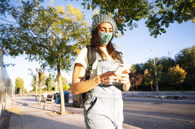 Femme portant un masque protecteur en utilisant un téléphone intelligent tout en marchant sur le sentier pendant la journée ensoleillée — Photo de stock