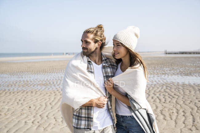 Jeune couple réfléchi couvert de couverture regardant loin tout en se tenant à la plage par une journée ensoleillée — Photo de stock