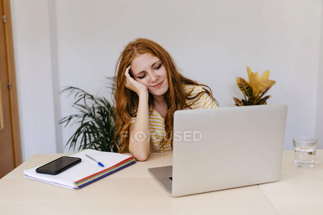 Mujer joven con la mano en el pelo usando el ordenador portátil mientras está sentado en casa - foto de stock