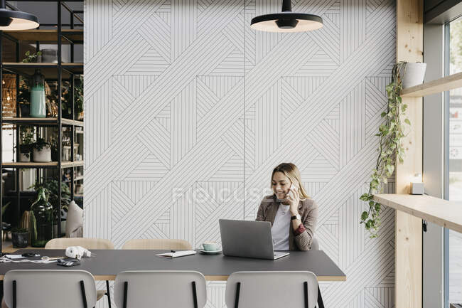 Lächelnde junge Frau, die mit dem Laptop im Café telefoniert — Stockfoto