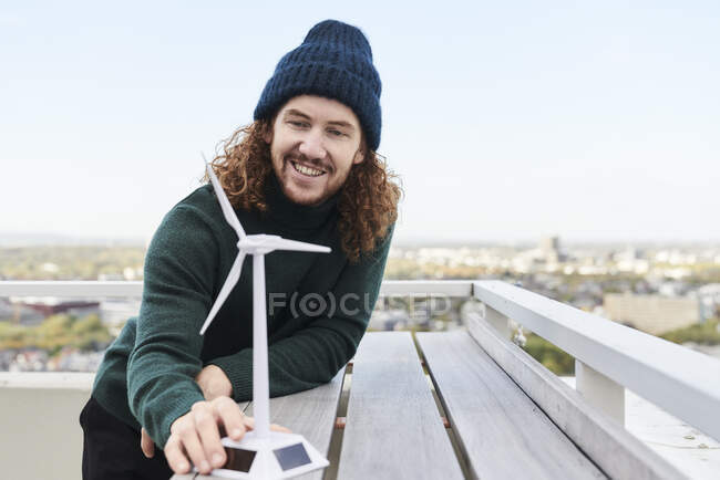 Smiling male hipster with windmill model on building terrace against sky — Foto stock
