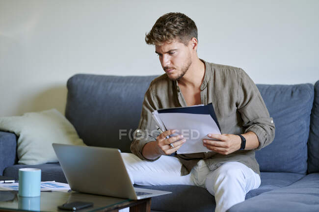 Businessman working on laptop while holding paper in living room — Stock Photo