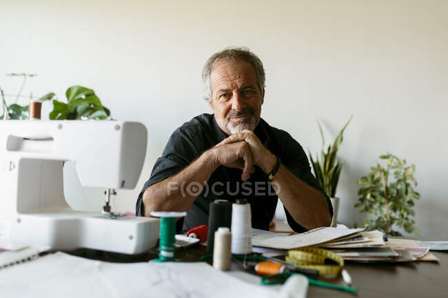 Tailleur masculin souriant assis à table contre le mur dans le studio de travail — Photo de stock