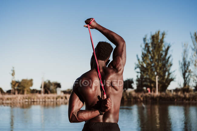 Giovane atleta maschio tirando banda di resistenza mentre guarda il mare contro il cielo limpido — Foto stock