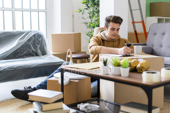 Young man writing on paper over cardboard box in new apartment — Stock Photo
