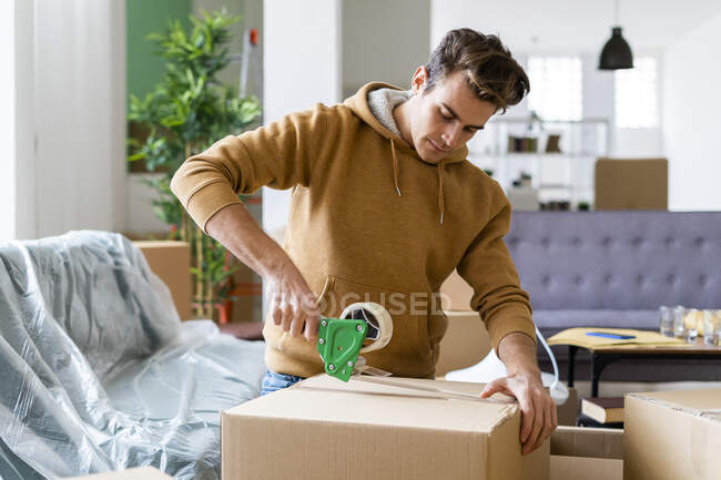 Young man applying adhesive tape on cardboard box while moving in new home — Stock Photo