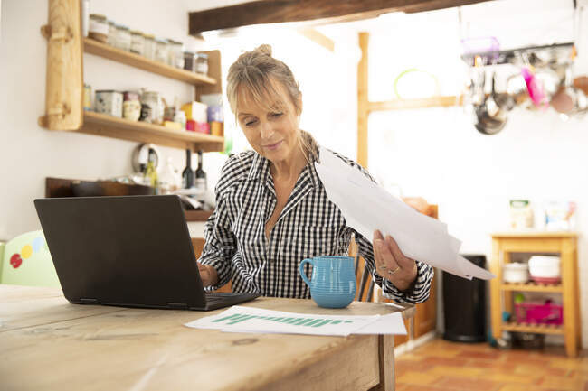 Senior woman working on laptop holding paper while sitting in kitchen at home — Stock Photo