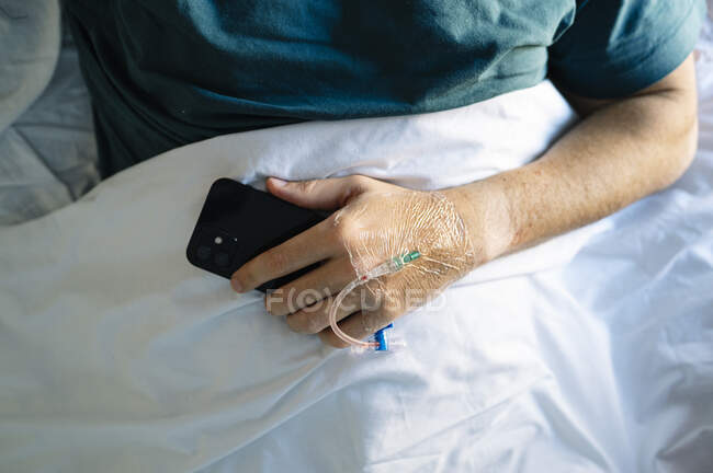 Patient tenant un téléphone portable avec perfusion intraveineuse couché sur le lit à l'hôpital — Photo de stock