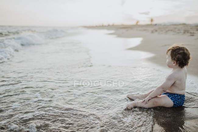 Carino bambino guardando la vista mentre seduto in acqua in spiaggia durante il tramonto — Foto stock