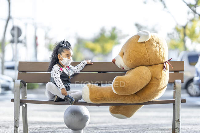 Дівчина з масками на обличчі бавиться з плюшевим ведмедиком, сидячи на відстані на лавці. — стокове фото