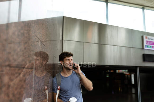 Hombre sonriente hablando a través del teléfono inteligente mientras mira hacia otro lado y se apoya en la pared de baldosas marrones en la estación de metro - foto de stock