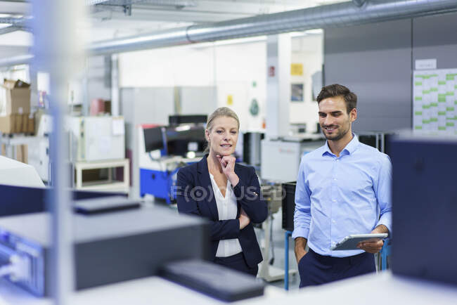 Sonrientes colegas profesionales discutiendo mientras miran la maquinaria en la fábrica iluminada - foto de stock