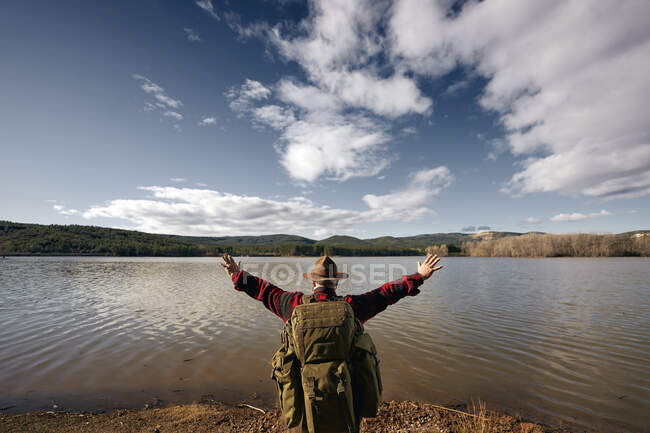 Bushcrafter mirando al lago con los brazos extendidos mientras está de pie en la naturaleza - foto de stock