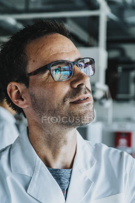 Científico usando anteojos mirando hacia otro lado mientras está de pie con su compañero de trabajo en el fondo en el laboratorio - foto de stock