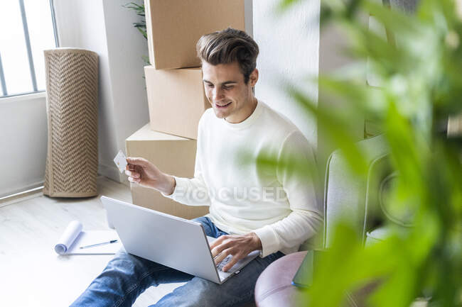 Smiling young man with credit card using laptop while moving in new home — Stock Photo