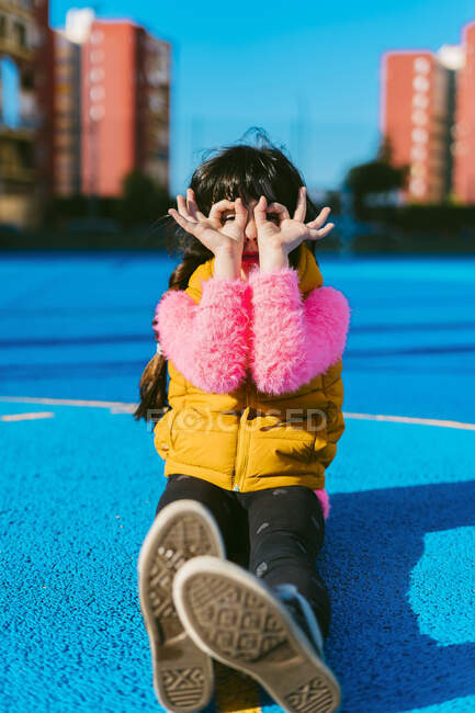Chica formando prismáticos con los dedos mientras se sienta en la cancha deportiva durante el día soleado - foto de stock