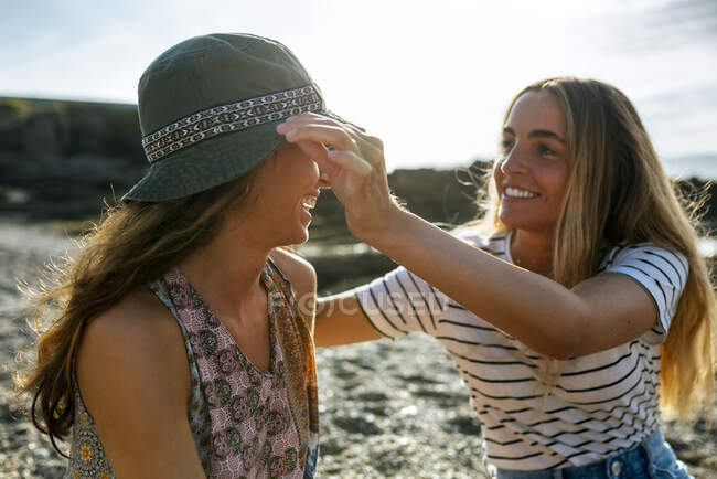 Smiling young woman adjusting hat of sister while enjoying weekend at beach — Stock Photo