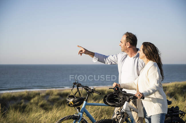 Couple heureux avec des vélos regardant la vue tout en se tenant sur la plage contre un ciel clair — Photo de stock