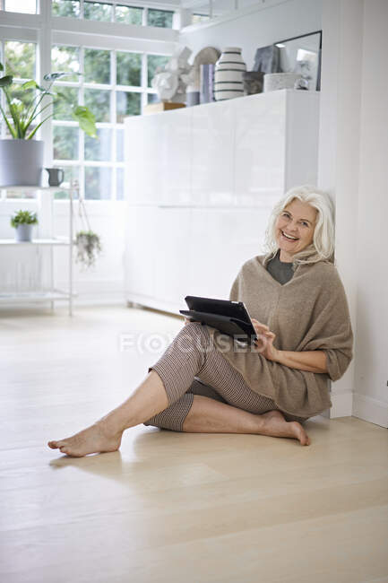 Mujer jubilada sonriente sentada en el piso con tableta digital en la sala de estar en el apartamento - foto de stock