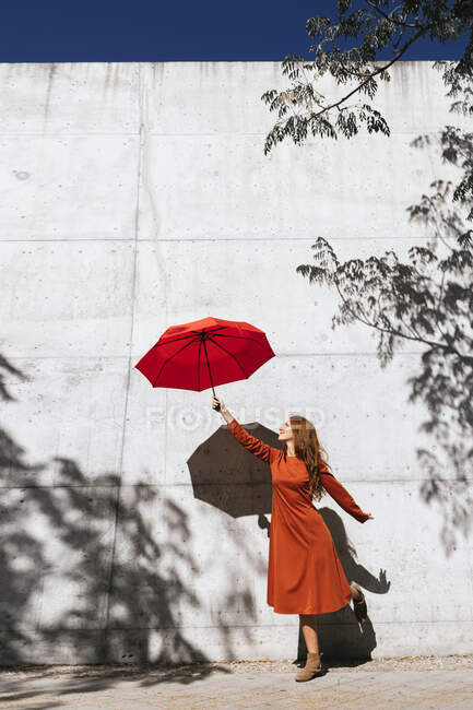 Mujer joven en vestido rojo sosteniendo paraguas volador mientras está de pie contra la pared de sombra de árbol - foto de stock