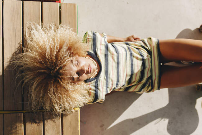 Donna con acconciatura bionda riccia con gli occhi chiusi sdraiata su sedile in legno durante la giornata di sole — Foto stock