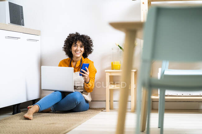 Mulher sorridente com laptop e telefone celular sentado no chão na cozinha em casa — Fotografia de Stock