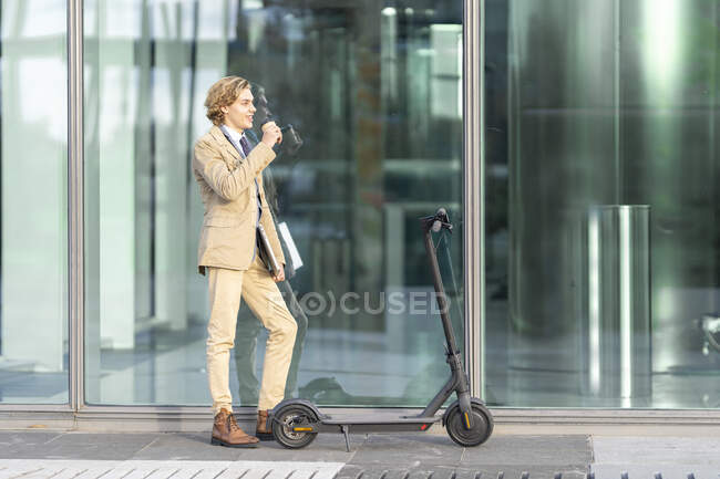 Бізнесмен з ноутбуком п'є каву, стоячи за допомогою електричного пускового скутера на скляній стіні — стокове фото