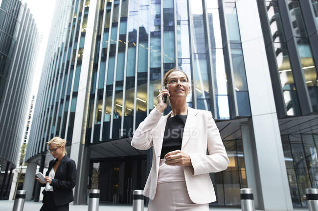 Female professional talking on mobile phone while standing near businesswoman against modern office building — Stock Photo