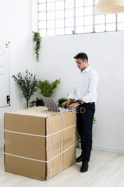 Jovem empresário usando laptop enquanto está em pé por caixa de papelão no local de trabalho criativo — Fotografia de Stock