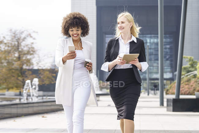 Smiling businesswomen walking with smart phone and digital tablet on footpath — Stock Photo