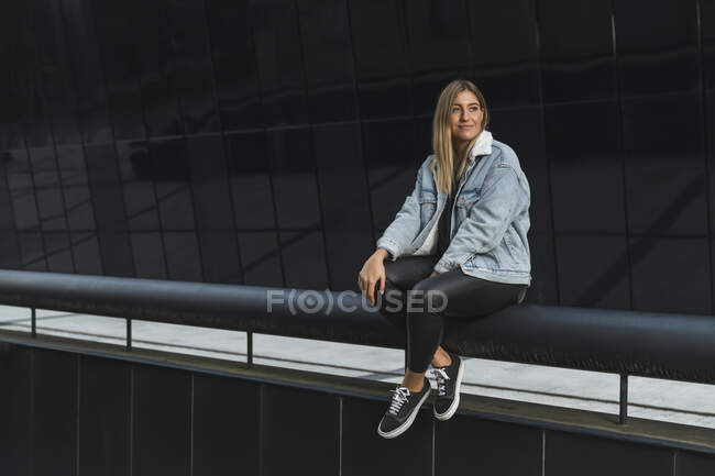 Giovane donna premurosa seduta su ringhiera contro l'edificio di vetro moderno — Foto stock