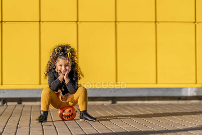 Chica sentada con la cabeza en las manos en maceta de Halloween contra la pared de patrón de cuadros amarillos - foto de stock