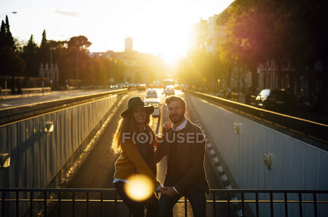 Couple holding hands while sitting on bridge railing together at city during sunset — Photo de stock