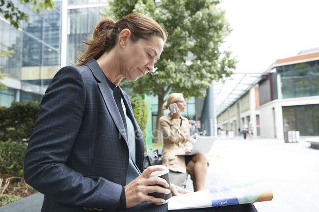 Empresarias leyendo papel mientras su colega habla en el teléfono móvil y el uso de la computadora portátil en segundo plano en la ciudad - foto de stock