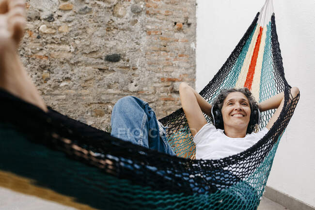 Mulher sorridente ouvindo música através de fones de ouvido enquanto relaxa na rede contra a parede no quintal — Fotografia de Stock