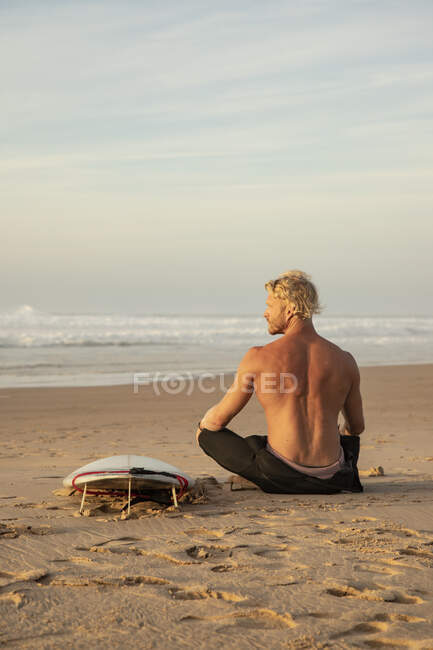 Surfista senza maglietta seduto a tavola da surf sulla sabbia mentre guarda il mare contro il cielo — Foto stock