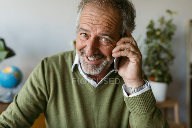 Homme mûr souriant parlant sur un téléphone portable tout en étant assis à la maison — Photo de stock